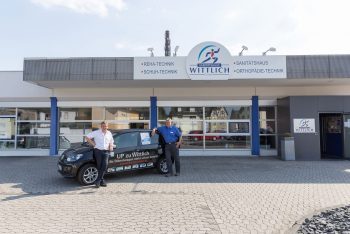 Drei Personen stehen neben einem Auto der Marke SH Wittlich vor einem Gebäude mit der Aufschrift Wittlich.
